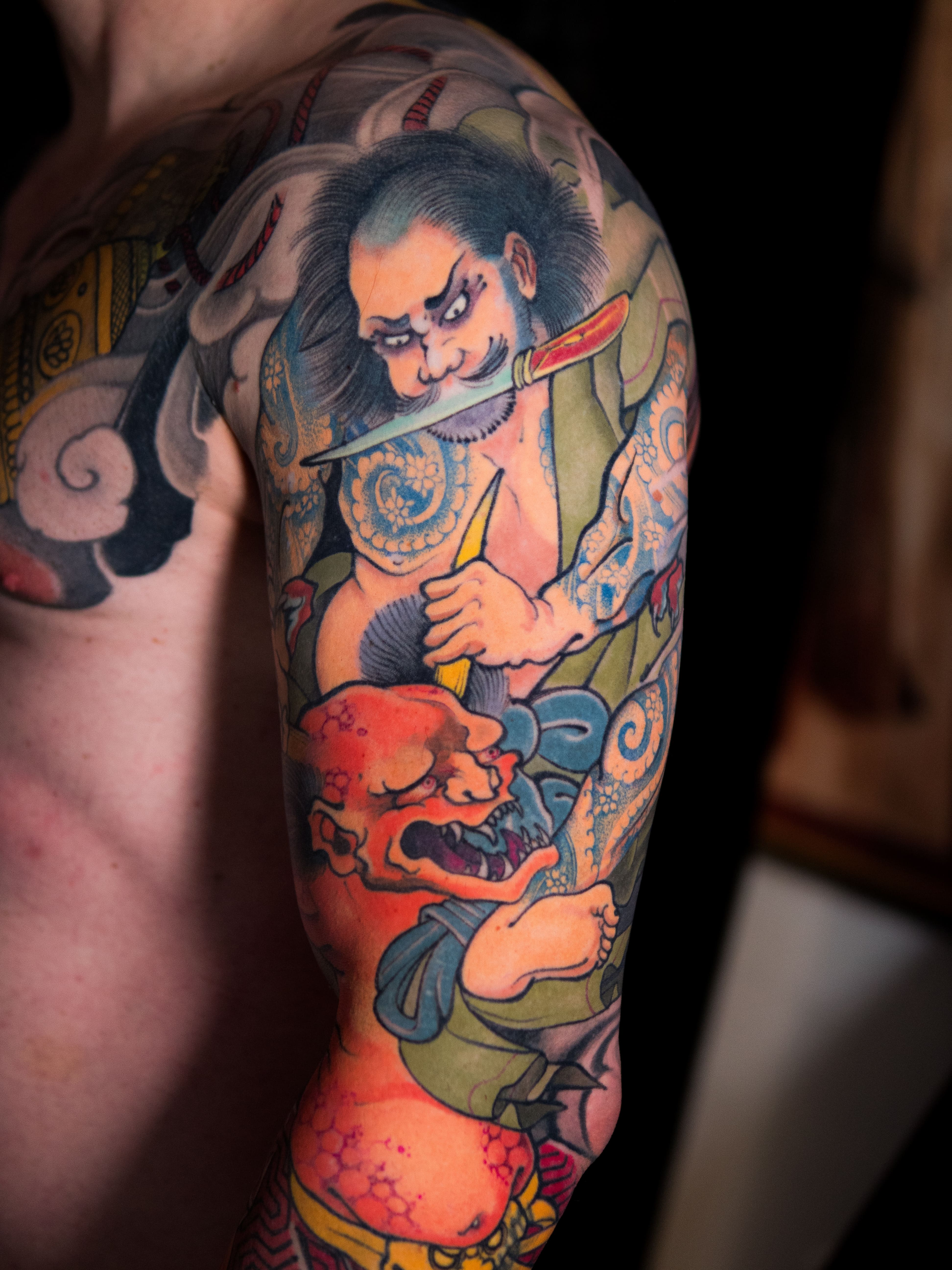 Na zdjęciu orientalny tatuaż, kolorowy, japoński, irezumi na ramieniu oraz przedramieniu przedstawiający wojownika z diabłem, wykonany w studio tatuażu Uroki w Warszawie